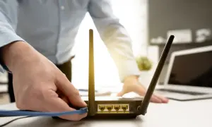 Dłoń podłącza kabel do routera, przygotowując reset. W tle widać laptopa.