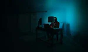 Osoba w masce hakerów siedzi przy biurku, korzystając z laptopa. W ciemnym pokoju, oświetlonym niebieskim światłem, przeglądarka darknet jest otwarta.