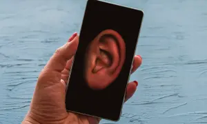 Dłoń trzyma smartfon, na którego ekranie widnieje ucho. Symbolizuje to szpiegowanie telefonu i podsłuchiwanie rozmów.