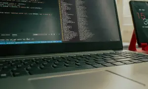 Laptop z otwartym terminalem i kodem, obok stoi smartfon na czerwonym stojaku.