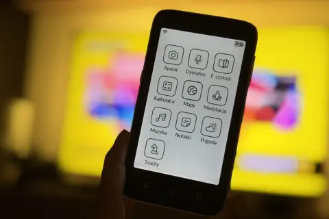Dłoń trzyma smartfon z ikonami aplikacji: aparat, dyktafon, e-czytnik, kalkulator, mapy, medytacja, muzyka, notatki, pogoda, szachy. To komunikator bez numeru telefonu.