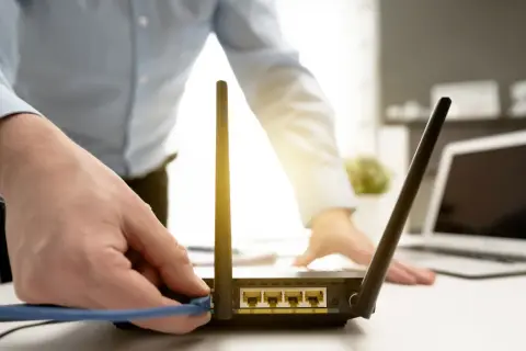 Dłoń podłącza kabel do routera, przygotowując reset. W tle widać laptopa.