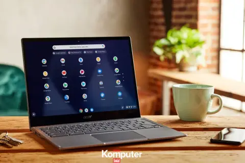 Laptop z systemem Chrome OS na drewnianym stole, obok kubek i telefon. Ikony aplikacji Google widoczne na ekranie.