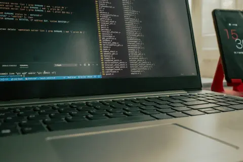 Laptop z otwartym terminalem i kodem, obok stoi smartfon na czerwonym stojaku.