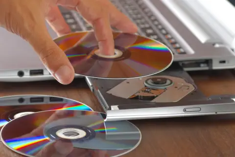Dłoń wkłada płytę do napędu laptopa, przygotowując się do nagrania danych. Program do wypalania płyt jest kluczowy do archiwizacji.