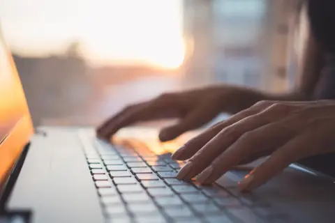 Dłonie piszą na klawiaturze laptopa z systemem MX Linux, w tle zachodzące słońce.