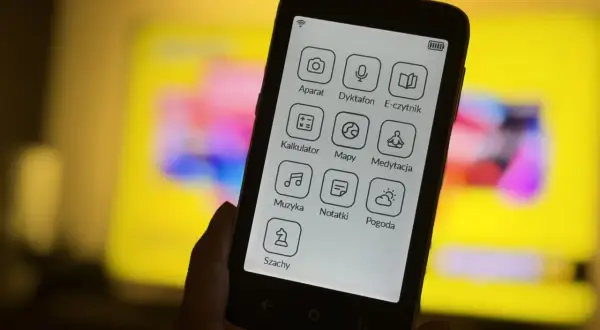 Dłoń trzyma smartfon z ikonami aplikacji: aparat, dyktafon, e-czytnik, kalkulator, mapy, medytacja, muzyka, notatki, pogoda, szachy. To komunikator bez numeru telefonu.
