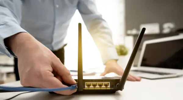 Dłoń podłącza kabel do routera, przygotowując reset. W tle widać laptopa.