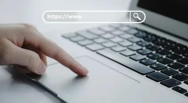 Dłoń na touchpadzie laptopa, pasek adresu z "https://www.", symbol lupy. Bezpieczna poczta online.