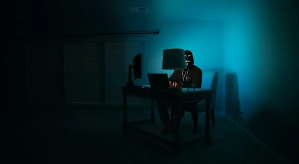 Osoba w masce hakerów siedzi przy biurku, korzystając z laptopa. W ciemnym pokoju, oświetlonym niebieskim światłem, przeglądarka darknet jest otwarta.