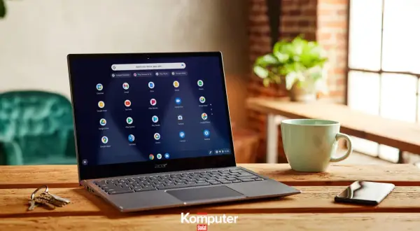 Laptop z systemem Chrome OS na drewnianym stole, obok kubek i telefon. Ikony aplikacji Google widoczne na ekranie.
