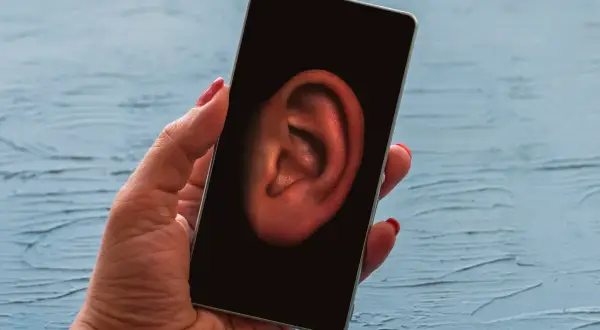 Dłoń trzyma smartfon, na którego ekranie widnieje ucho. Symbolizuje to szpiegowanie telefonu i podsłuchiwanie rozmów.