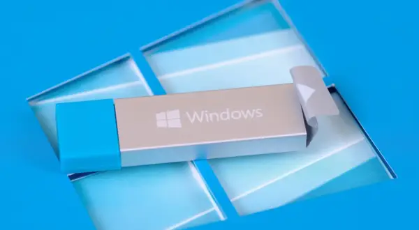 Srebrny pendrive startowy Windows 10 z niebieską nakładką, leżący na niebieskim tle z logo Windows.