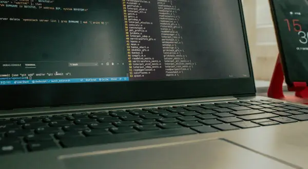 Laptop z otwartym terminalem i kodem, obok stoi smartfon na czerwonym stojaku.