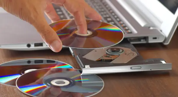 Dłoń wkłada płytę do napędu laptopa, przygotowując się do nagrania danych. Program do wypalania płyt jest kluczowy do archiwizacji.