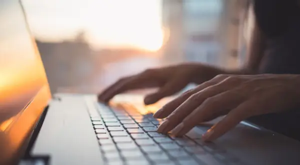 Dłonie piszą na klawiaturze laptopa z systemem MX Linux, w tle zachodzące słońce.