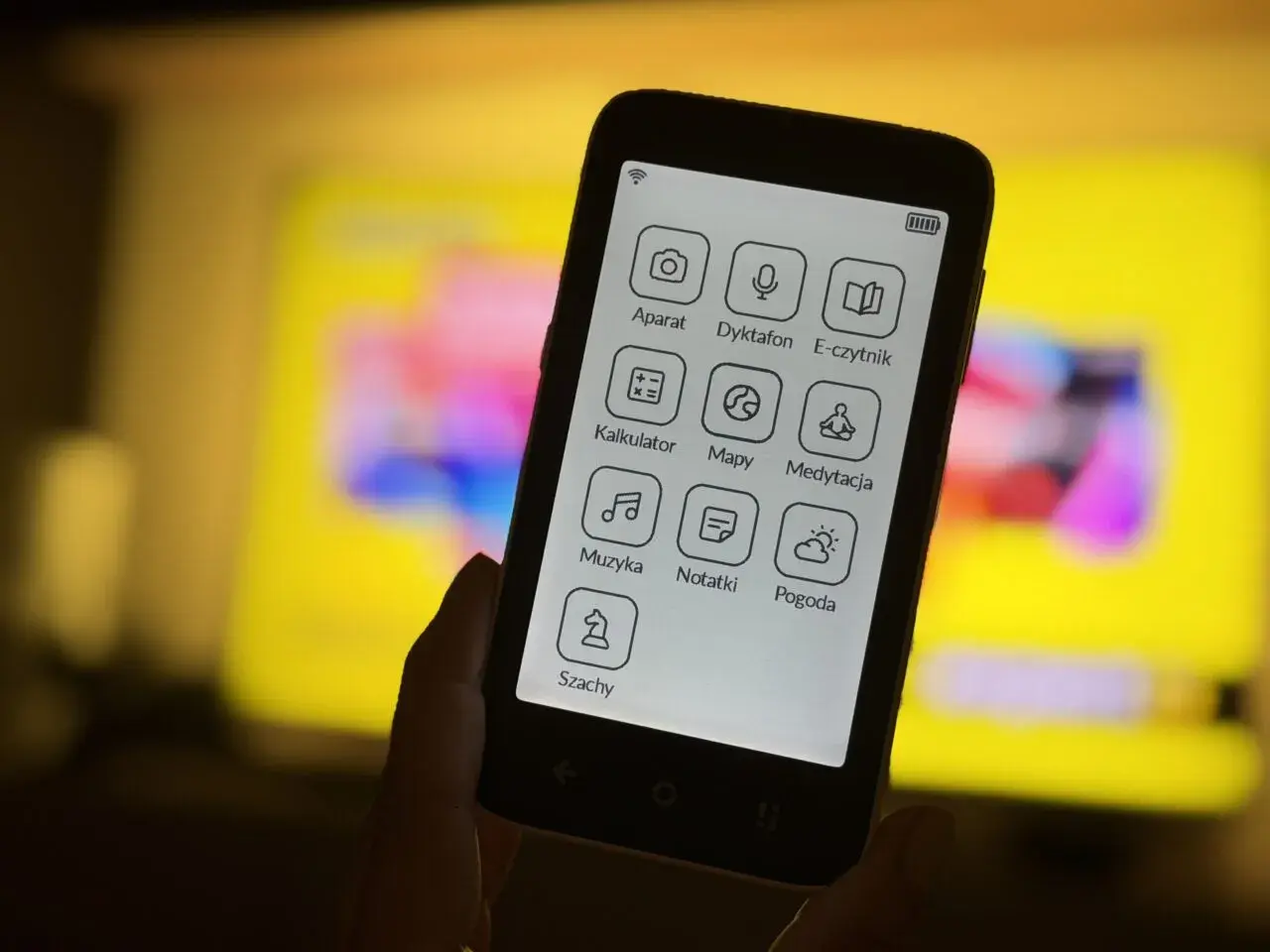 Dłoń trzyma smartfon z ikonami aplikacji: aparat, dyktafon, e-czytnik, kalkulator, mapy, medytacja, muzyka, notatki, pogoda, szachy. To komunikator bez numeru telefonu.