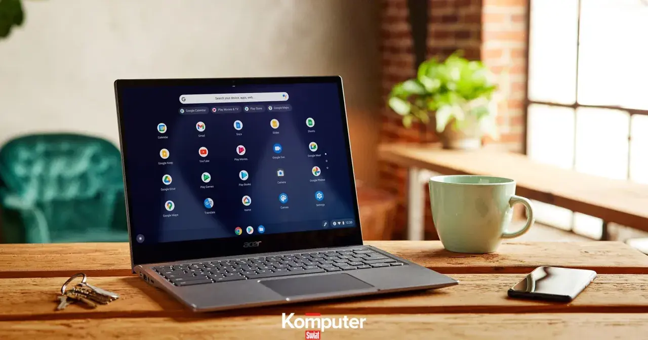 Laptop z systemem Chrome OS na drewnianym stole, obok kubek i telefon. Ikony aplikacji Google widoczne na ekranie.