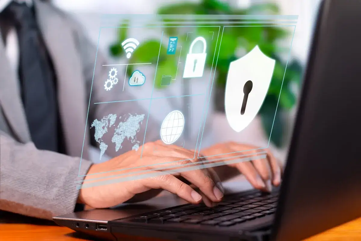 Osoba pracuje na laptopie, a przed nią wyświetla się interfejs z ikonami symbolizującymi bezpieczeństwo w internecie: globus, mapa świata, chmura, kłódka i tarcza.