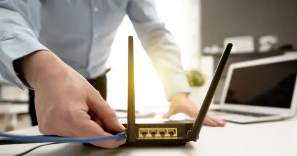 Dłoń podłącza kabel do routera, przygotowując reset. W tle widać laptopa.