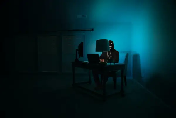 Osoba w masce hakerów siedzi przy biurku, korzystając z laptopa. W ciemnym pokoju, oświetlonym niebieskim światłem, przeglądarka darknet jest otwarta.