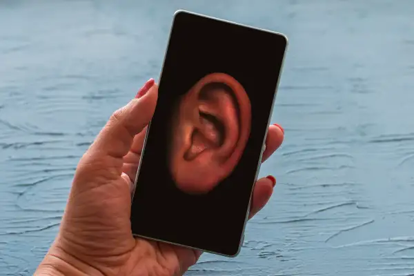 Dłoń trzyma smartfon, na którego ekranie widnieje ucho. Symbolizuje to szpiegowanie telefonu i podsłuchiwanie rozmów.